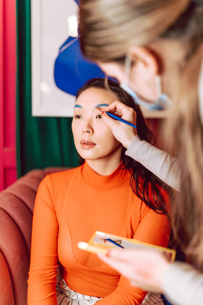 services-03 A makeup artist carefully applies eye makeup to an Asian woman seated in a modern indoor setting.