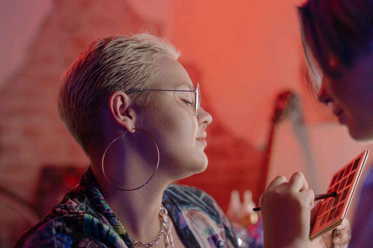 A woman with eyeglasses and hoop earrings enjoys a makeup session indoors.