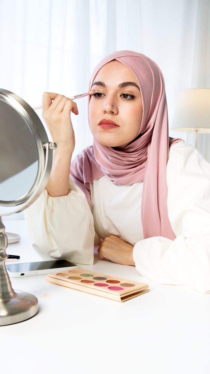 services-04 Middle Eastern woman wearing a hijab applies eyeshadow using a brush indoors.